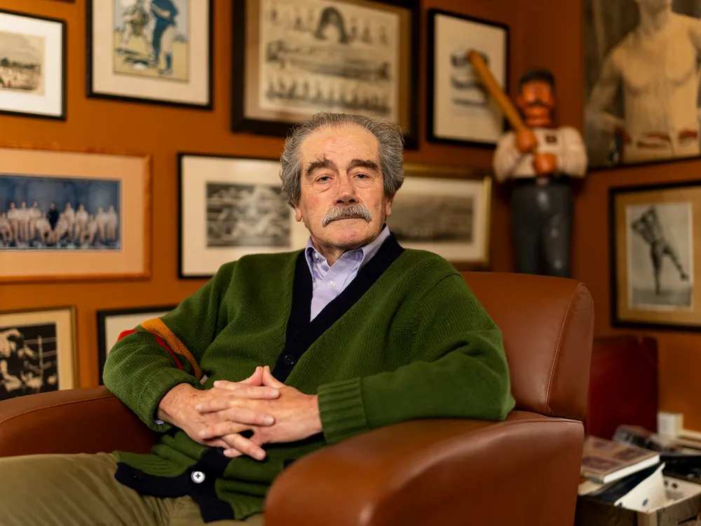 A man in a green sweater sits in front of a wall of hanging photographs