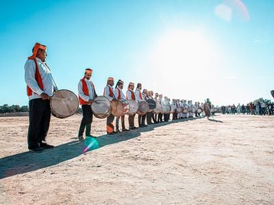 A line of drummers wearing red vests and white shirts stand in the dessert on a clear, blue-skied day.