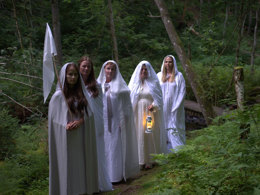 Five adults dressed in white dresses, robes, and veils pose along a forest path. One carries a lit lantern, and another carries a white flag.