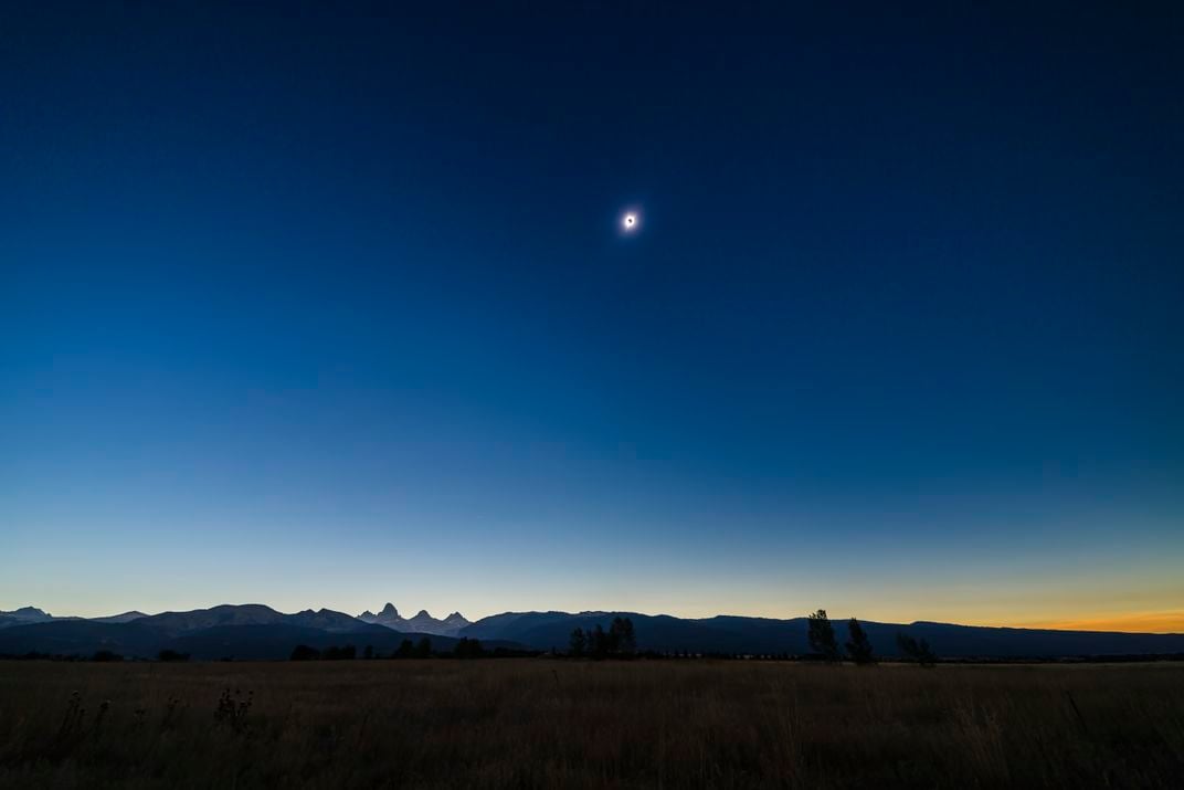 The sun during a total eclipse, seen small in the sky with the moon blocking out its center, over a dark sky with sunset colors along the horizon